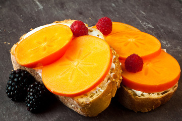 Fototapeta na wymiar Sliced persimmon fruit with cream cheese on freshly baked corn bread. Served with fresh raspberries and blackberries on a stone slate table top.