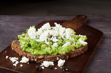 Healthy snack of dark rye bread topped with avocado and crumbled feta cheese served on a stone slate table top.