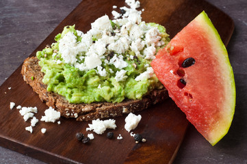 Healthy snack of dark rye bread topped with avocado and crumbled feta cheese served with a slice of fresh water melon.