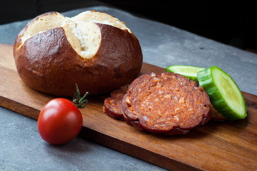 Rustic rough cut salami served with fresh pretzel bread with tomato and cucumber. Served on a wooden plank on a slate stone table.