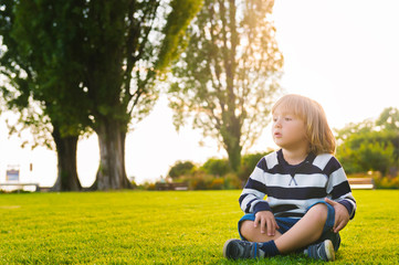 Portrait of beautiful smiling cute little boy. 3-4 years old little child playing outside in green summer or spring park. Boy sitting alone on green grass at sunset, wearing stripe jersey sweatshirt