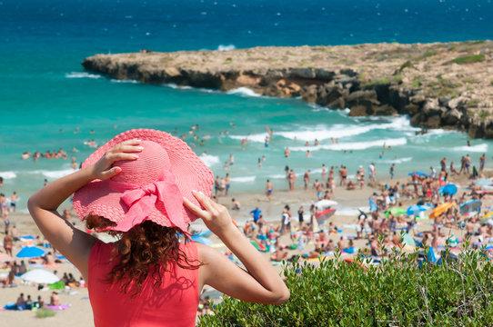 Tourist girl with a pink hat looking at the crowded beach of Calamosche, Sicily, from a panoramic point