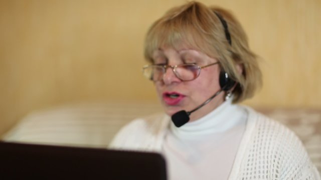 Woman communicates via laptop. Woman with headphones with microphone talks to a customer via computer. Female with notebook and headset with a microphone. Defocused video at the beginning