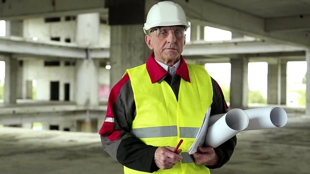 Master builder at project site. Chief architect in white hard hat, protective goggles and building drawings at construction site. Builder looks at the camera. Worker with design drawings