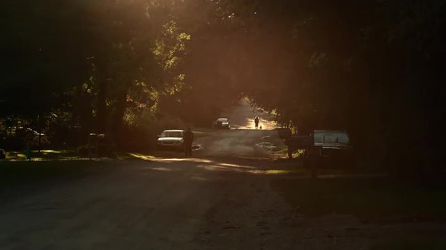 Sun setting over a dirt road in the Mid West, USA.  Lens flares through locked-off shot.  People walking in the distance. Originally recorded in 4K, UHD.
