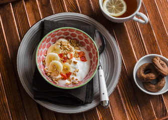 Yogurt with banana, dried fruit and granola, chocolate chip cookies and tea with lemon on brown wooden background. Delicious Breakfast