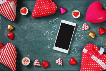 Fototapeta na wymiar top view image of smartphone, colorful heart shape chocolates, fabric hearts on blackboard background. valentine's day celebration concept 