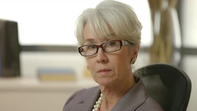 An older female CEO or senior executive turns slowly and looks intently into the camera.