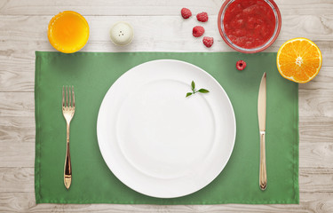 Empty plate on the table waiting for serving. Top view of tablecloth, jar of jam, juice, raspberries, orange.