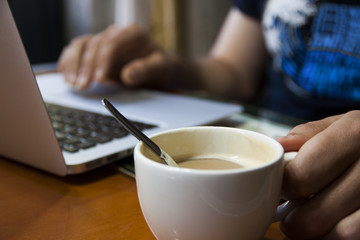 chico moreno con barba tomando el desayuno, un café con leche y trabajando con su ordenador en la cocina de su casa