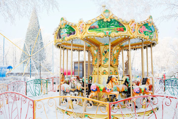 Merry-go-round traditional horses covered with snow. Behind the carousel big Christmas tree. 