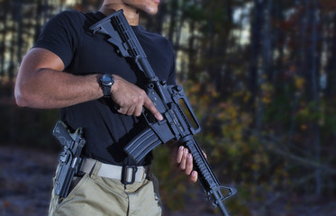 Man in a forest holding an assault rifle with handgun on his side ready to shoot