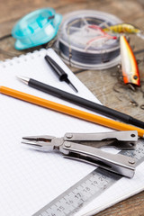 writing-book with fishing tackles and design tools on wooden boa