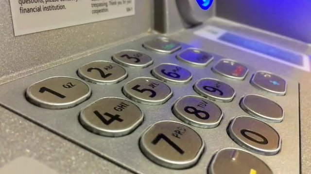 Blue light flashes from automated teller machine behind brushed metal number key pad buttons.