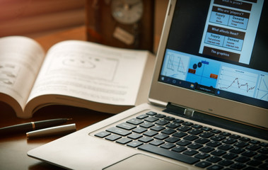  Laptop with diagrams , book , pen and  clock at office desk. Shallow depth of field. Warm light balance. 