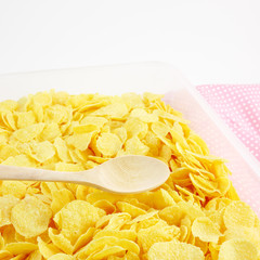 The close up of tasty golden corn flakes and wooden spoon in plastic container box on pink cotton fabric.