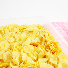 The close up of tasty golden corn flakes in plastic container box on pink cotton fabric.