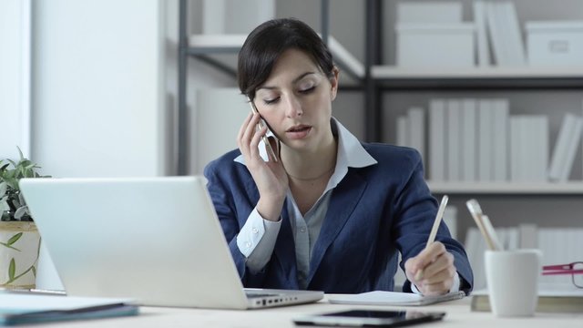 Businesswoman working at office desk, she is talking on the phone and writing down notes on a notebook