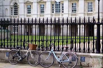 Two women's town bicycles, facing each other, leaning against an ornamental iron railing in front of a historic university building in Cambridge, England
