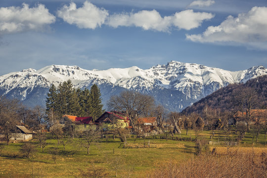 Spring sunny countryside landscape with snowy Bucegi mountains and traditional Romanian houses and farmland in Predelut village, near Bran, Transylvania region, Romania.