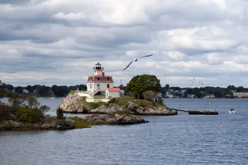 Poster Sun illuminating Pomham Rocks lighthouse after stormy weather in Rhode Island as seagull flies in front of the beacon. Sun breaking through rain clouds.