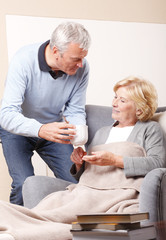 Obraz premium Portrait of lovely senior couple relaxing at home. Elderly woman sitting in armchair at living room with her husband. Old man holding in hand a glass of water while assisting his wife.