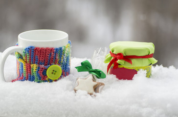 Jar of jam in the snow with a biscuit and a cup in the knit cover