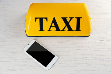 Transportation concept. Yellow taxi sign with smart phone on white wooden background, close up