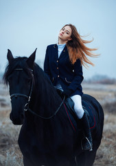 Obraz premium a tall girl with long blond hair in a jockey outfit with a beautiful black horse in an empty snow-covered field in winter