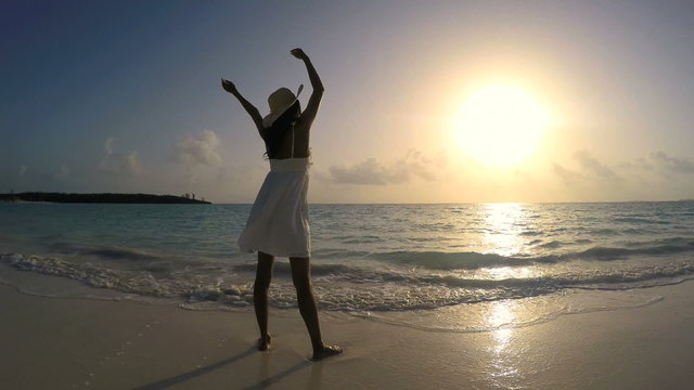 Young ethnic female in white dress dancing on sunrise beach
