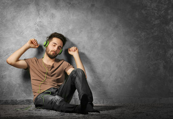 Young bearded man listens music with headphones sitting on grey wall background