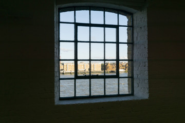 View framed by black surrounding wall to recent architectural buildings on other side River Thames through old wharf warehouse building windows from the dark interior at Wapping, London buildings 