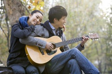 A young man plays the guitar as he girlfriend leans on him affectionately