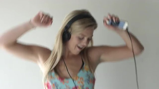 Tilt up from feet to the face of a young blonde woman in blue dress, smiling and dancing while wearing headphones connected to a cellphone playing music.  Medium close up.  Recorded in 4K, UHD.