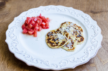 Delicious ricotta pancakes or fritters with parmesan cheese and chopped red tomatoes salad with olive oil on patterned plate over stylized old aged wooden table background close-up, copy space
