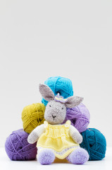 A knitted, soft toy rabbit  sitting in front of six balls of wool and isolated on a white background.