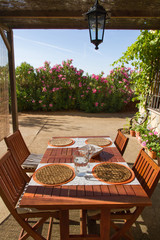 Mesa en Portico Exterior de una Casa con alguna Vajilla Puesta preparada para cuatro comensales, con sillas, en el jardin exterior de una casa.  Pan, jarra y copa con agua. Rodeada de azaleas