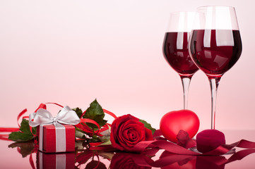 Valentine's decoration with rose, wine glasses and gift box. Glossy red table top. Background slightly lit by red.