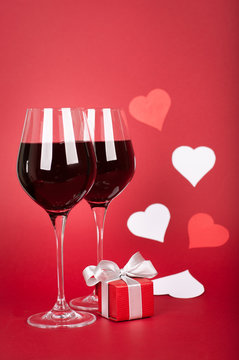 Valentine’s day - wine glasses, hearts and elegant gift box. Vertical position. Red matte background. White and red elements.