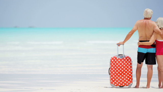 Barefoot Mature Caucasian Couple In Swimwear On A Beach With A Suitcase