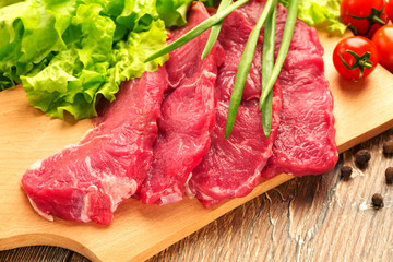 Fresh, crude stakes from meat veal on a wooden chopping board with setion, black pepper, lettuce leaves, cherry tomatoes and green onions