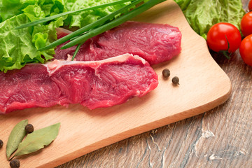 Fresh, crude stakes from meat veal on a wooden chopping board with setion,black pepper, lettuce leaves,fennel, bay leaf, green onions and cherry tomatoes