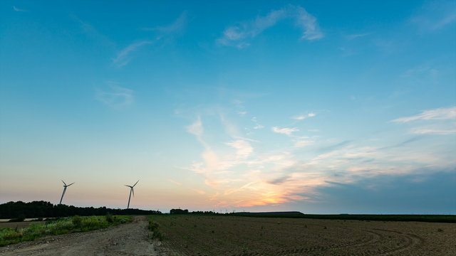 Time lapse sequence of rotating wind turbines during a sunset with a dirt road leading into the image in 4K.