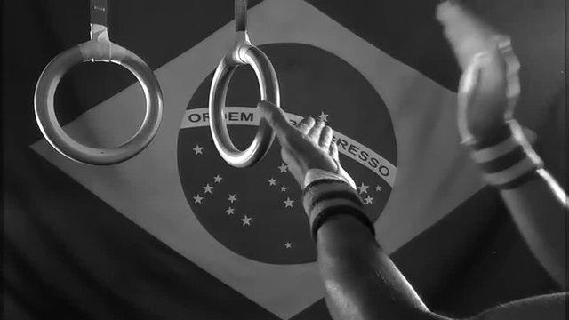 Taped hands of gymnast clapping white chalk powder into a cloud under gymnastic rings in front of a Brazil flag background in gritty black and white
