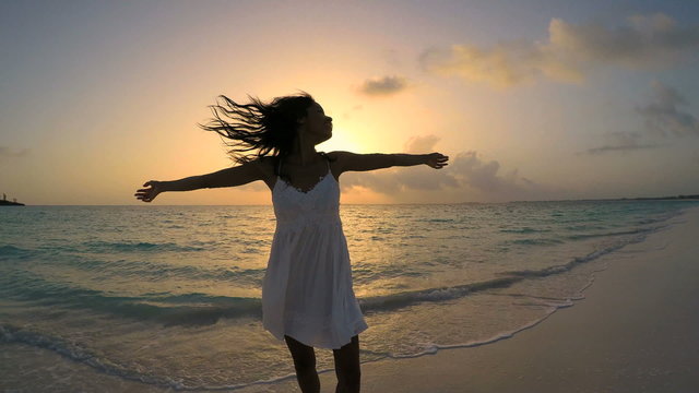 Young barefoot ethnic female in white dress dancing on sunset beach