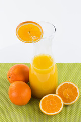 Frisch gepresster Orangensaft in einer Karaffe. Zwei ganze und halbierte Orangen um die Karaffe gelegt auf grünem Untergrund.