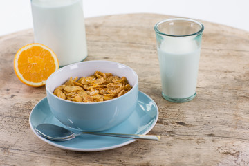 Müslifrühstück auf hellem Holztisch. Blaue Schale auf einem Teller mit Löffel. Ein Glas Milch und eine aufgeschnittene Orange.