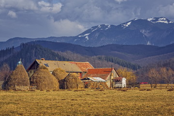 Rural scenery with traditional Romanian rustic farm with haystacks and grassland in the backyard, near Postavaru massif, Brasov county, Romania.