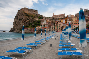 Blue umbrella and beachchair at  the beach in front of small italian town on cliff under blue sky