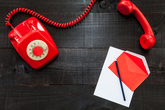 The image shows a vintage red phone on dark wood and paper,  pencil and a red envelope